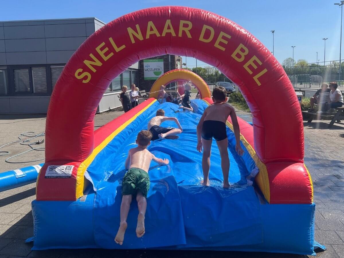 Kinderen racen over de buikschuifbaan naar de bel Kinderen glijden lachend over een buikschuifbaan tijdens een zomerfeest van Move for Fun en proberen als eerste de bel te raken.