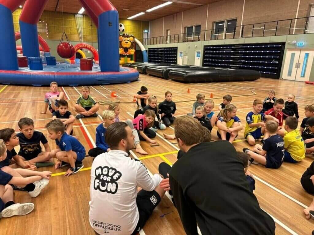 Kinderen luisteren naar uitleg tijdens een sportdag georganiseerd door Move for Fun in een sporthal met opblaasbare spellen.
