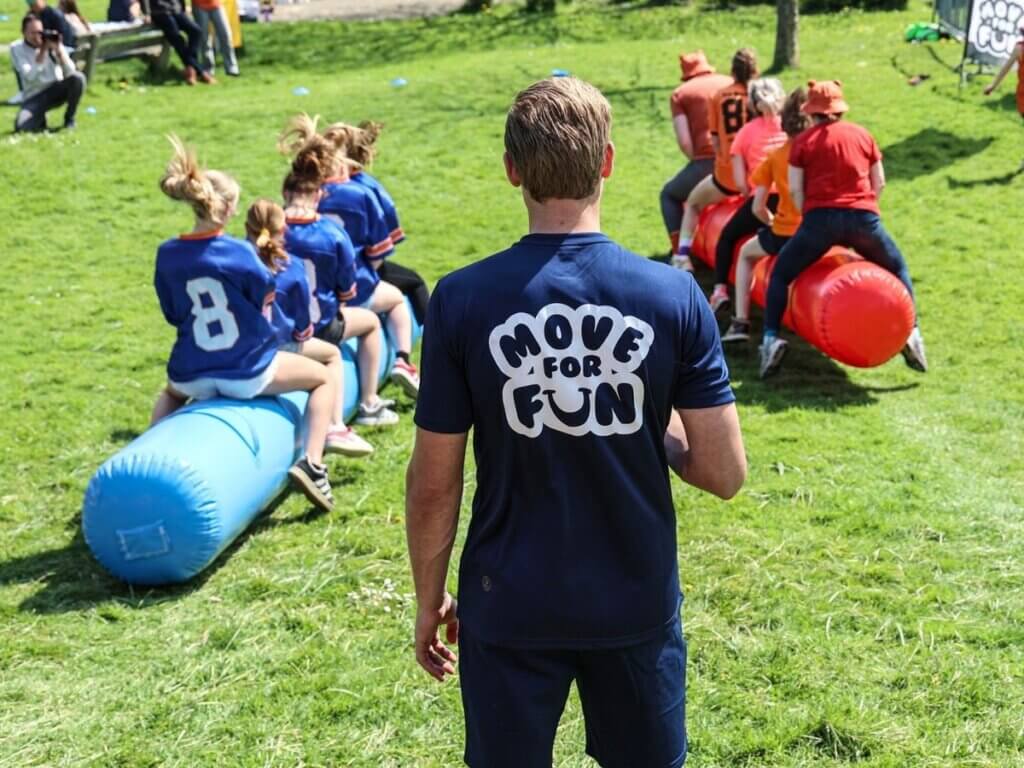 Begeleider van Move For Fun leidt een zeskampwedstrijd met teams op skippyslangen tijdens een sportieve buitendag.