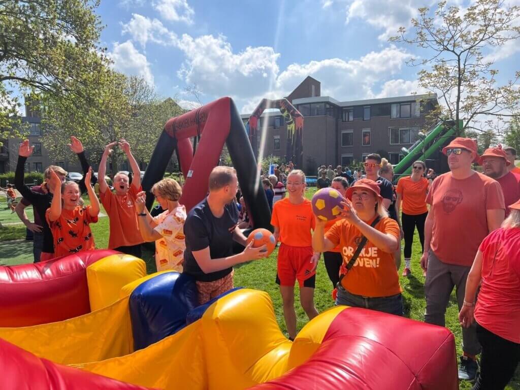 Groep mensen in oranje kleding die meedoen aan een Koningsdag groepsspel op een springkussen tijdens een zonnig evenement.