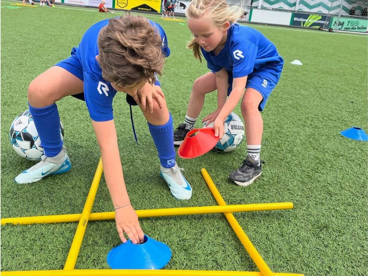 Reactiespel voetbal – Kinderen spelen een speelse trainingsvorm Twee kinderen pakken pionnen tijdens een reactiespel met bal op een voetbalveld.